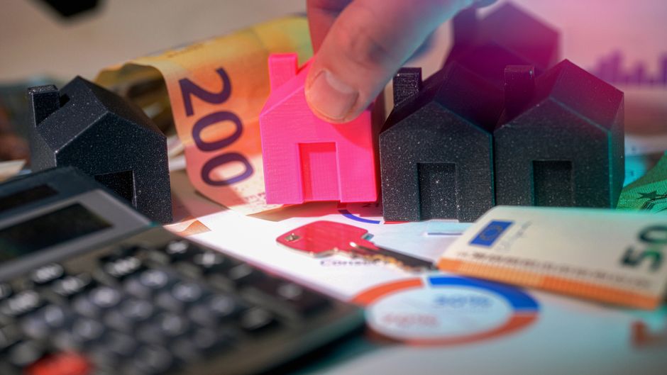 A close-up view of a hand holding a small, bright pink miniature house figure positioned among several dark grey or black miniature house models, all laid out on a surface covered with euro banknotes,