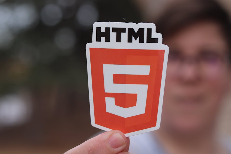 A person holding a sticker displaying the HTML5 logo with a white border, featuring a prominent orange shield shape with a stylized white number '5' in the center. The person's hand, with short, clean